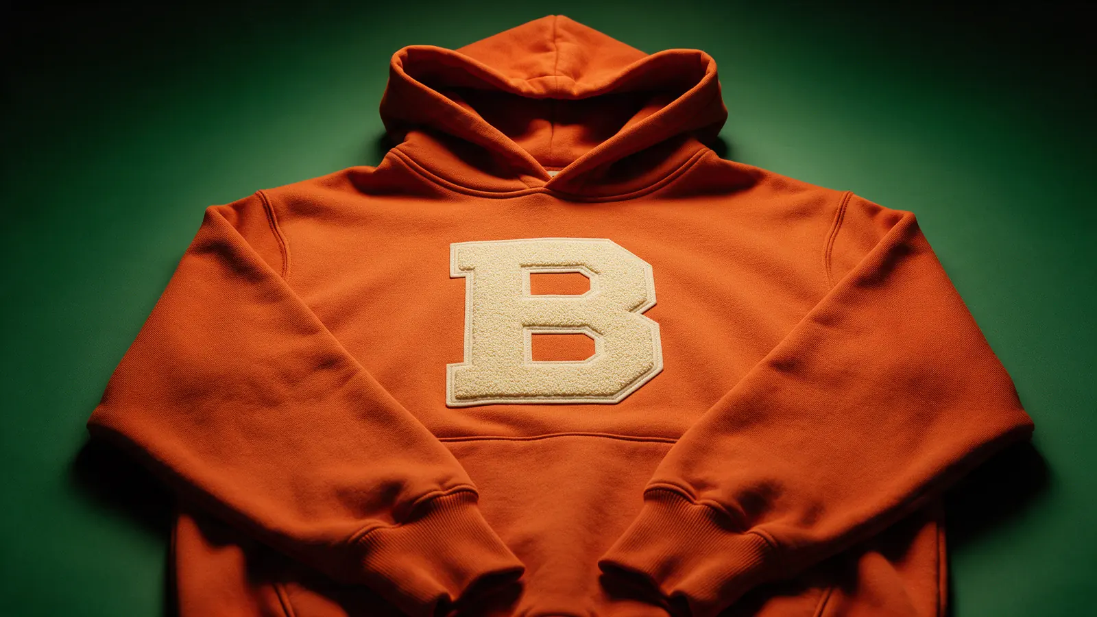 Hoodie color óxido con letra chenille crema sobre fondo verde